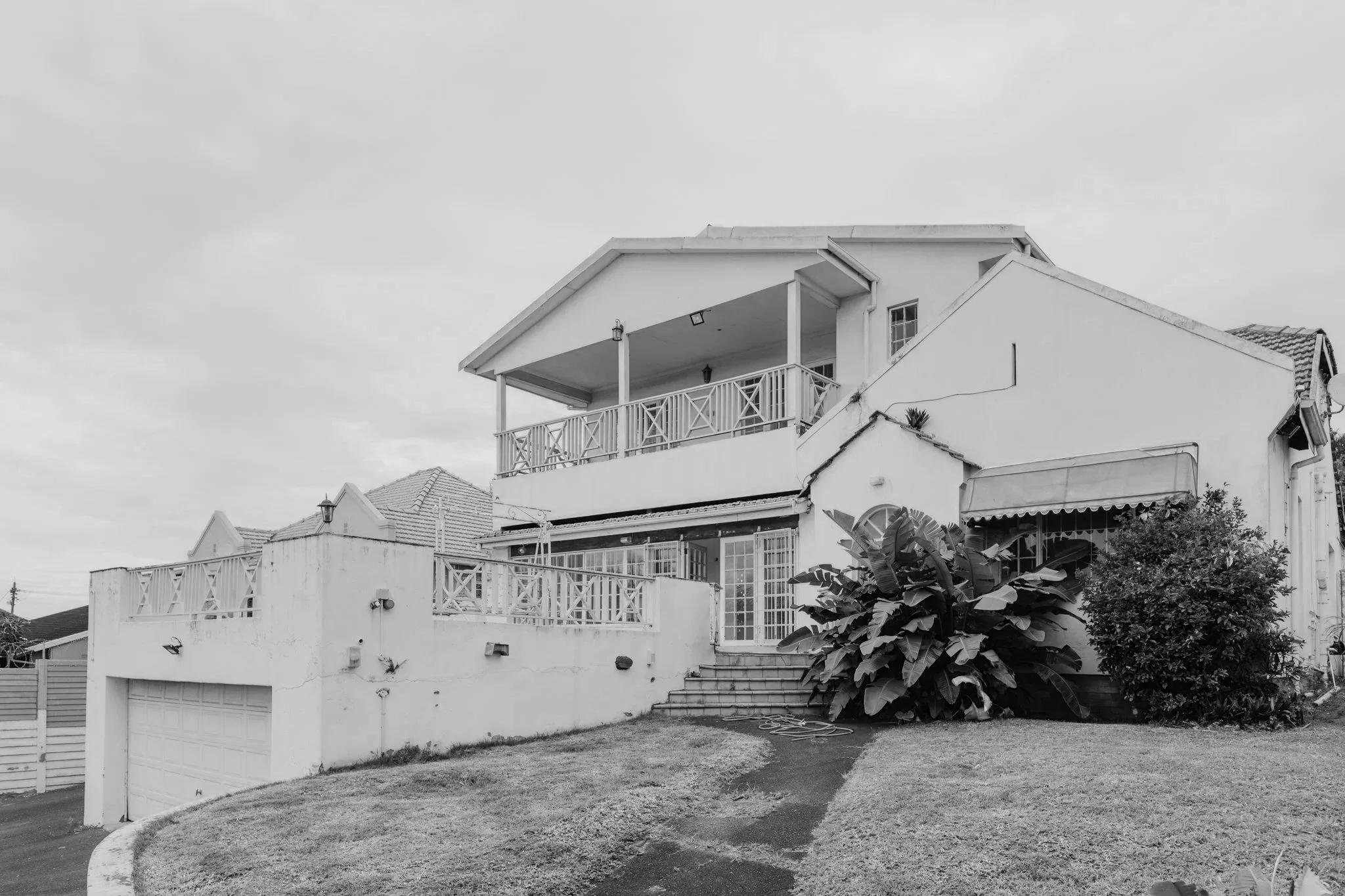 A two-story white house with a red tile roof, a front porch with stairs leading up to it, and a blue awning over one window. The house has a balcony and garden plants in front, on a cloudy day.