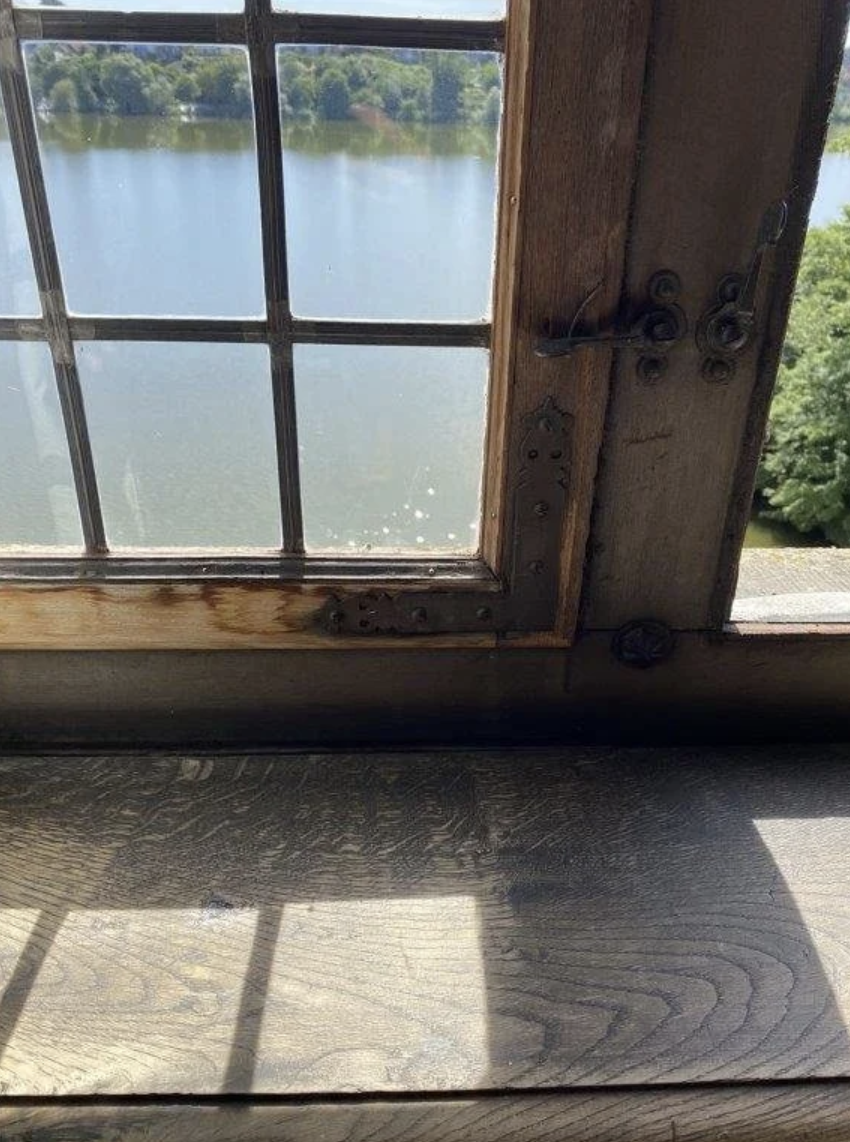 A wooden window with metal hinges and latch, showing a view of a body of water and trees outside, with sunlight casting shadows on the wooden interior floor.