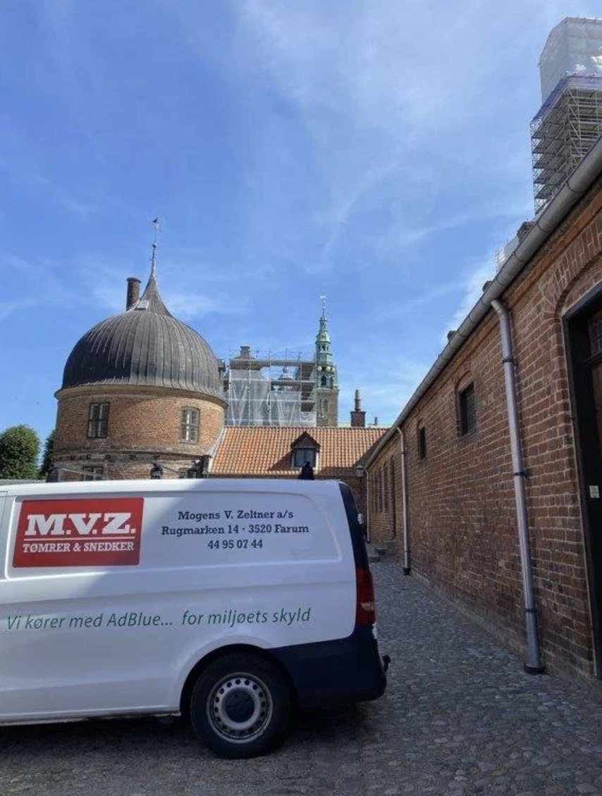A white company van parked in front of historic brick buildings with domed and spired rooftops, under a blue sky with some clouds.