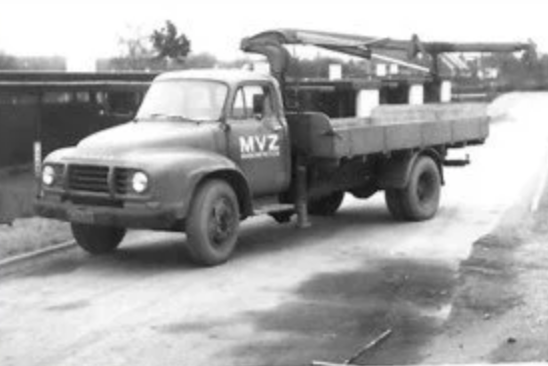 Black and white photo of a vintage flatbed truck with a mounted crane, parked on a dirt road.