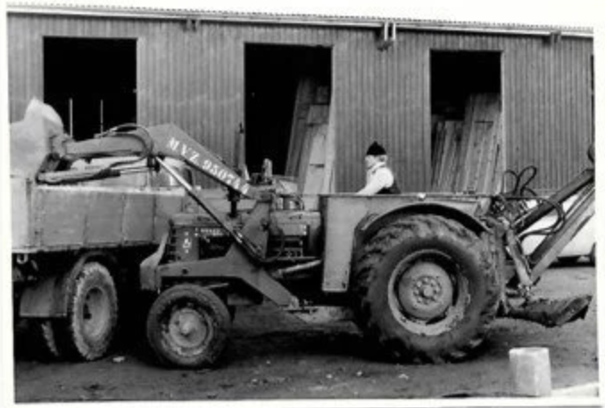 Black and white photo of a small tractor with a front loader attachment working in front of a wooden building with three gaps in the wall.