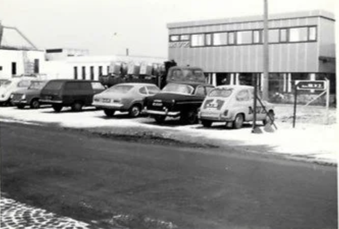 A black-and-white photo of a parking lot with several cars and a young child playing near a lamp post. There is a modern two-story building in the background with large windows.