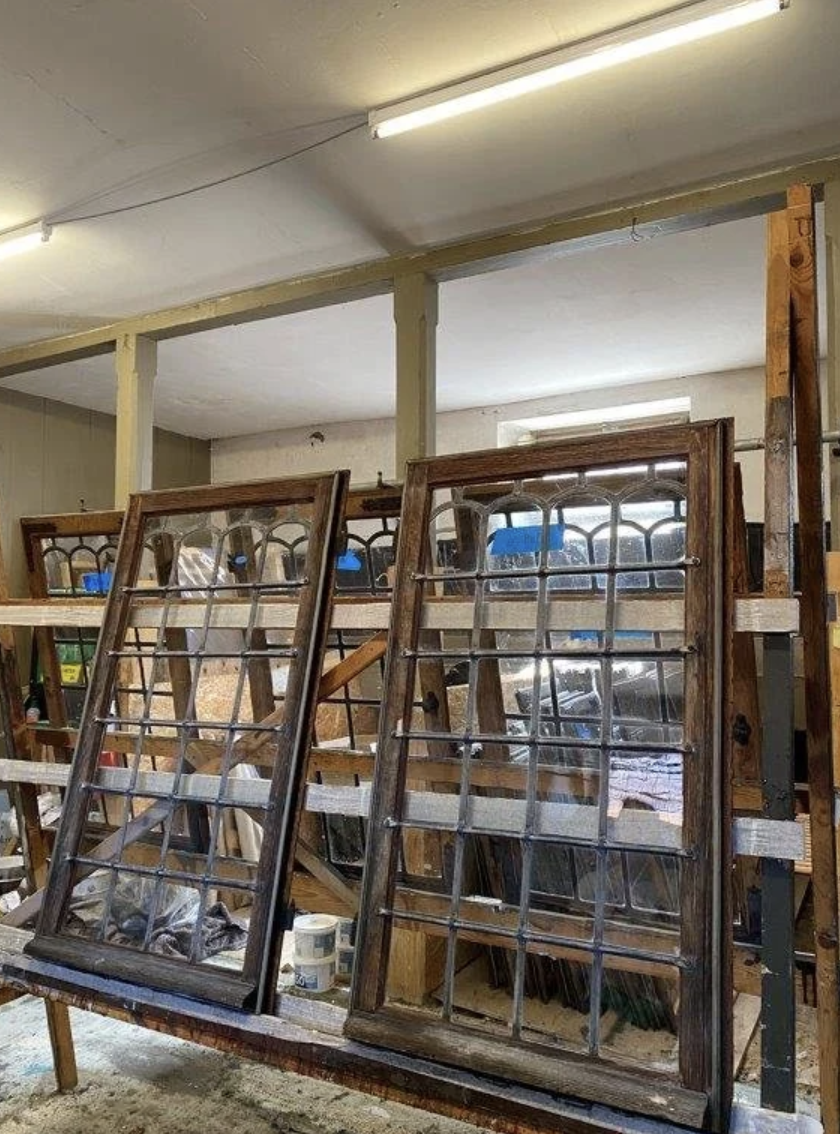 Two antique wooden window frames with glass panes leaning against a worktable in a workshop.