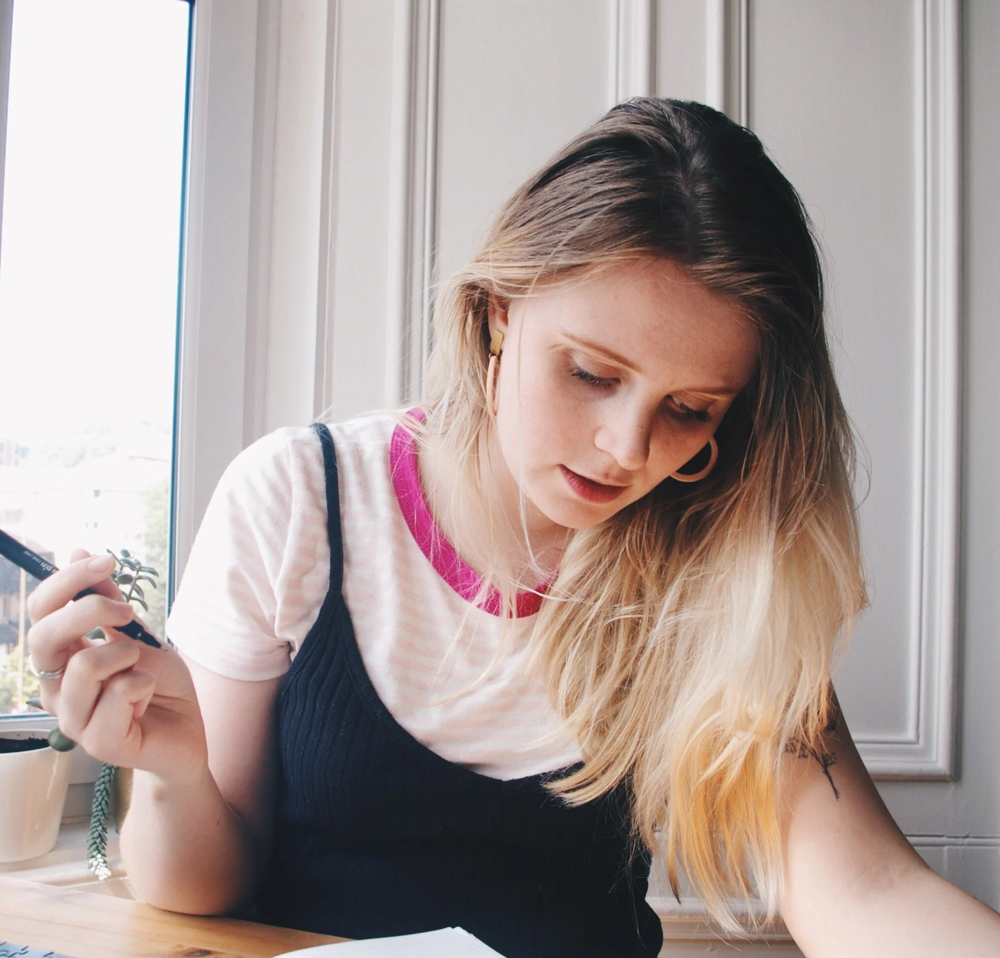 Amy with blonde hair and hoop earrings is sitting at a table near a window, holding a pen and looking down at some papers.