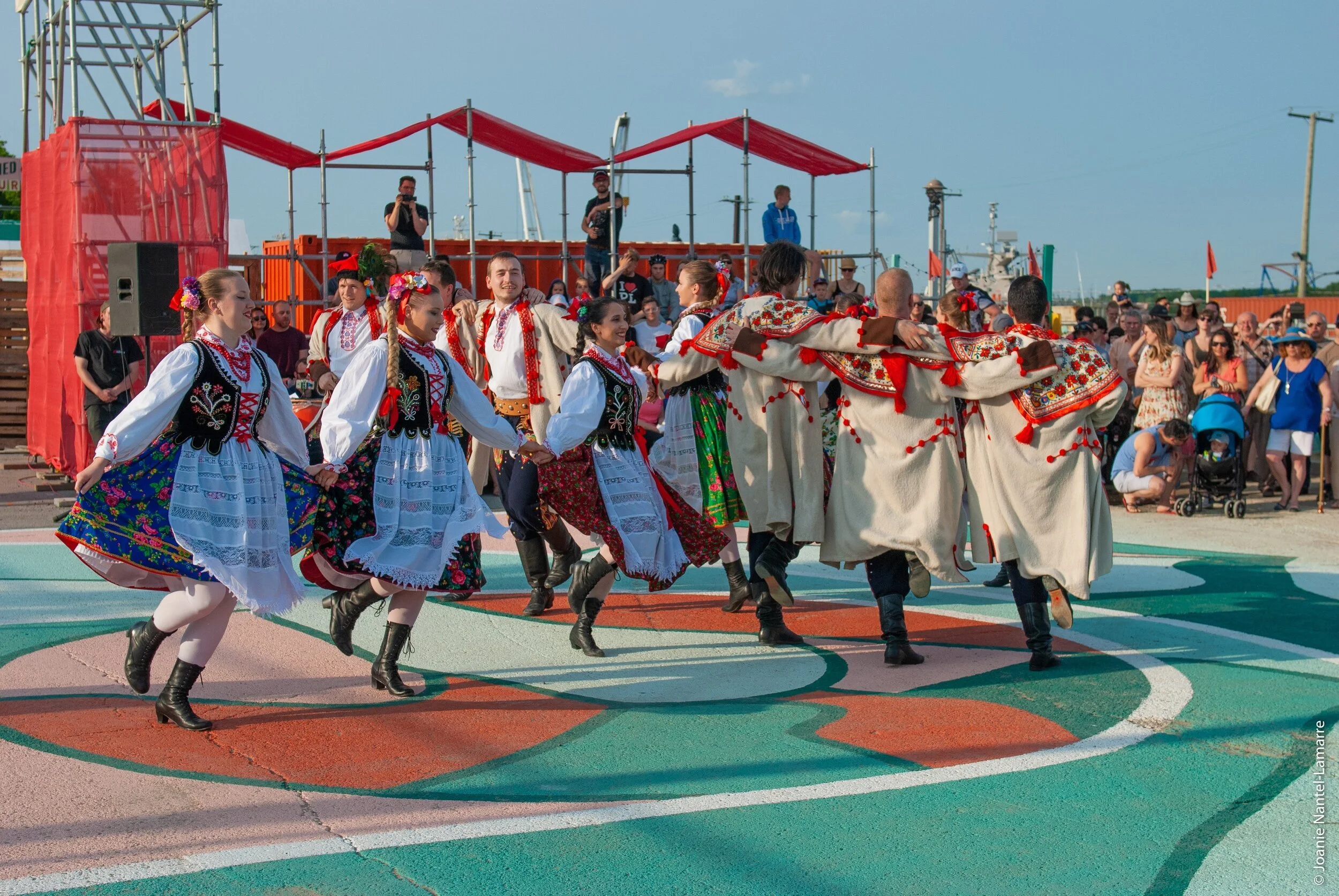 Group of people in traditional folk costumes dancing in a circle on an outdoor court during a cultural event. Spectators watch in the background, some taking photos, and there is a stage with red canopy in the distance.