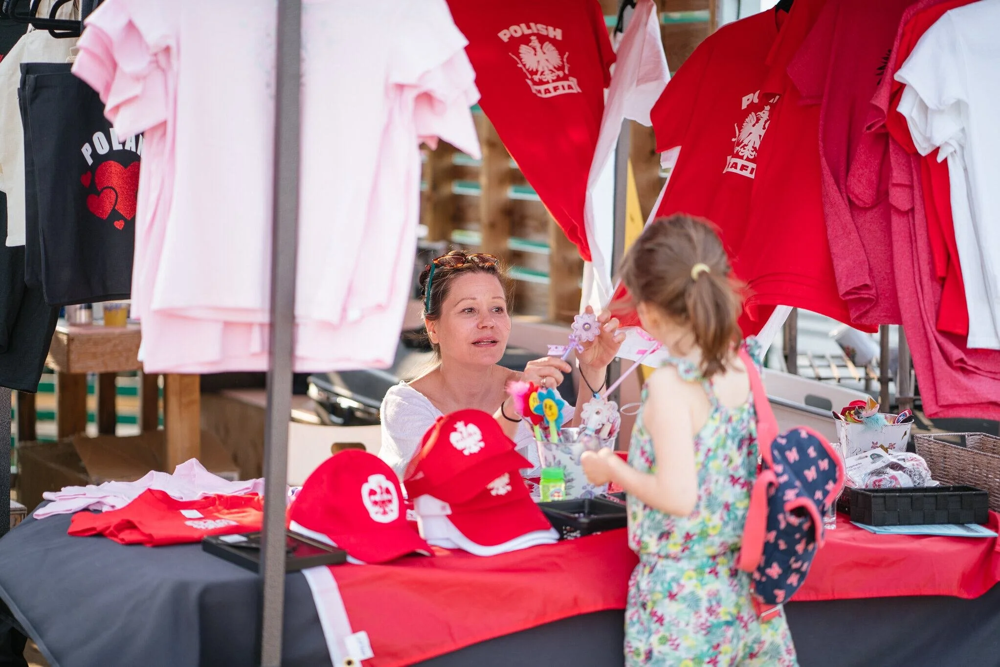 A woman selling red, white, and pink clothing and accessories at a stall with a young girl browsing. The clothing has Polish eagle emblem and Hearts, some labeled 'POLISH'.