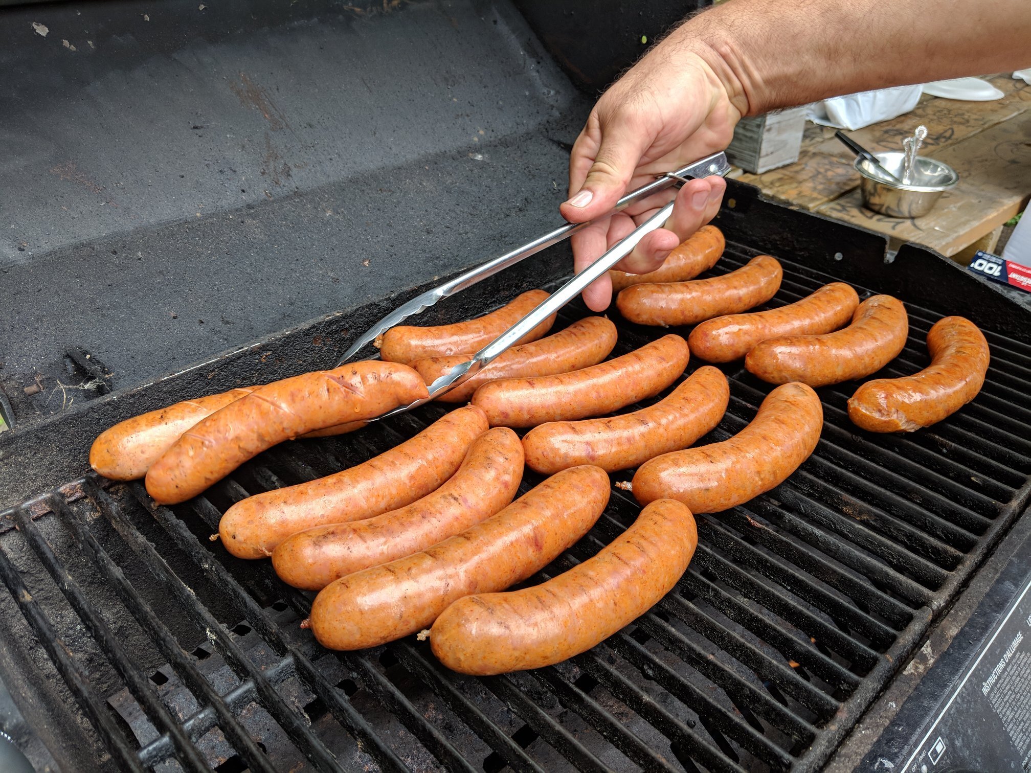 Several raw sausages being grilled on a barbecue grill, with a person's hand using tongs to turn them. In the background, there is a small bowl with some water and a spoon on a wooden surface.