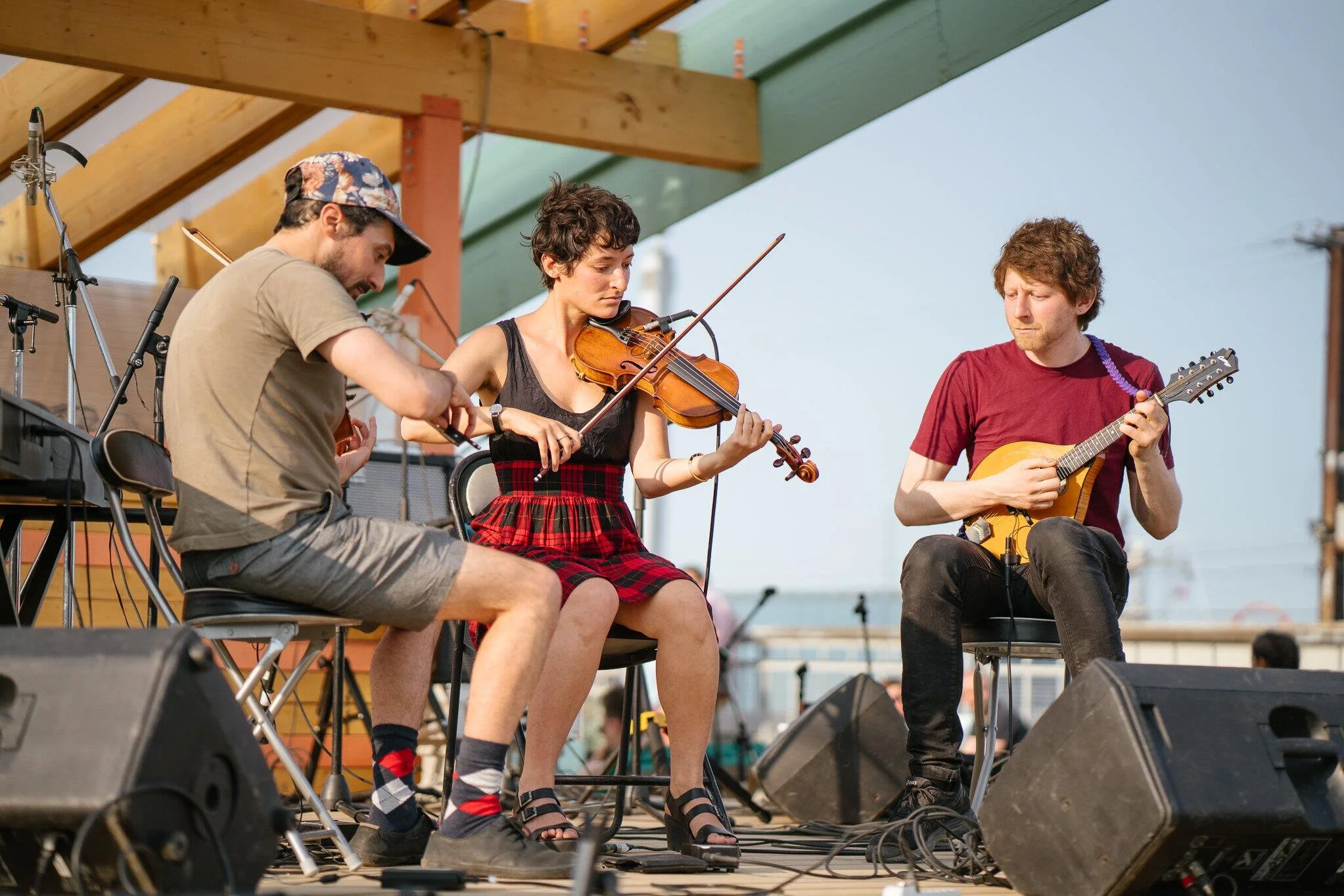 Three musicians performing on stage outdoors under a wooden roof. The woman in the center is playing a violin, and the man on the right is playing a guitar. The man on the left is adjusting equipment.