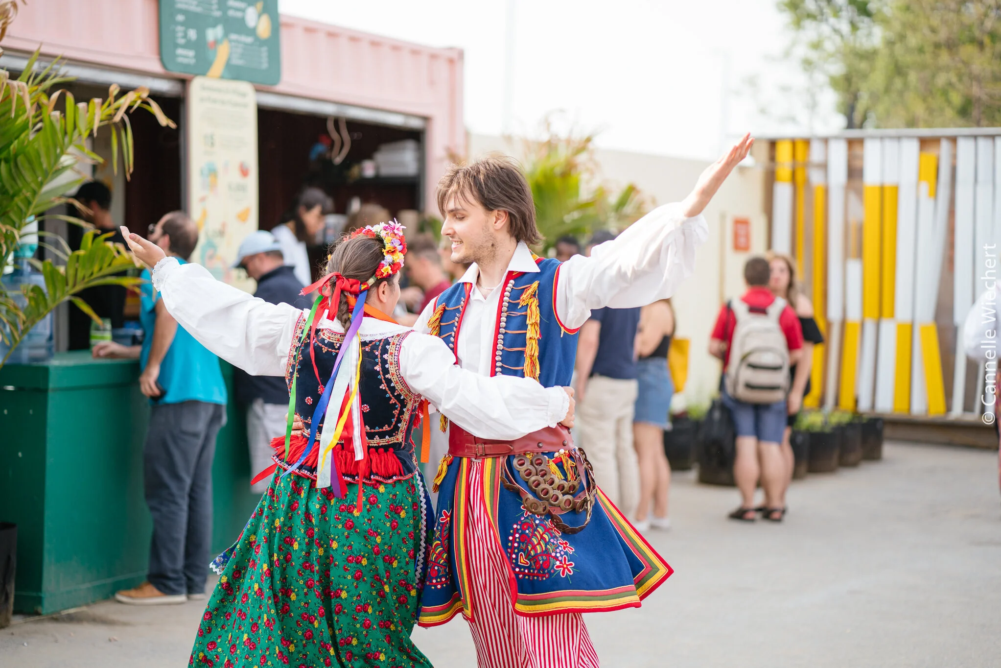 Two people in traditional folk costumes dancing during a cultural festival, with a food stand and people in the background.