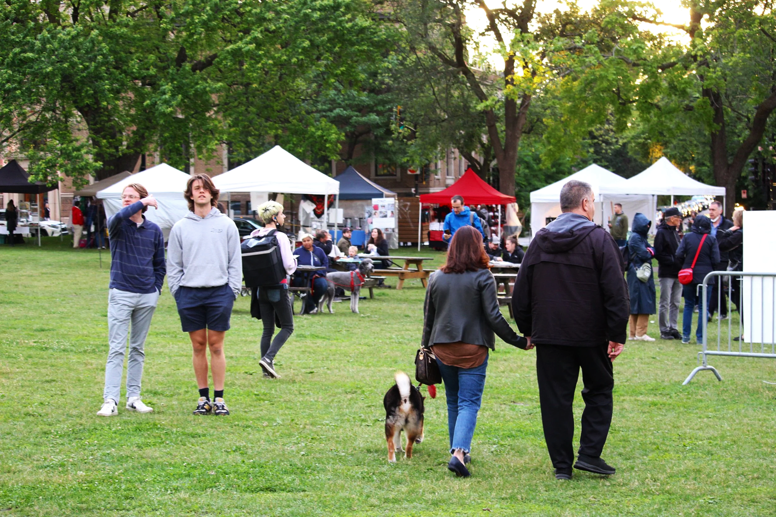 People walking and socializing in a park with food stalls and tents, green trees and grass, and overcast sky.