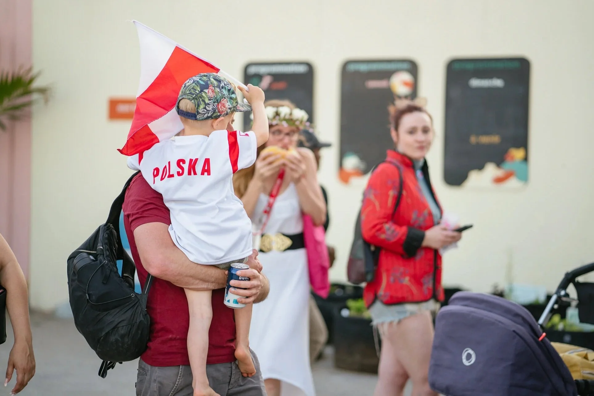 Child wearing a cap with a floral pattern holding a red and white flag and sitting on an adult's shoulders at a public gathering.
