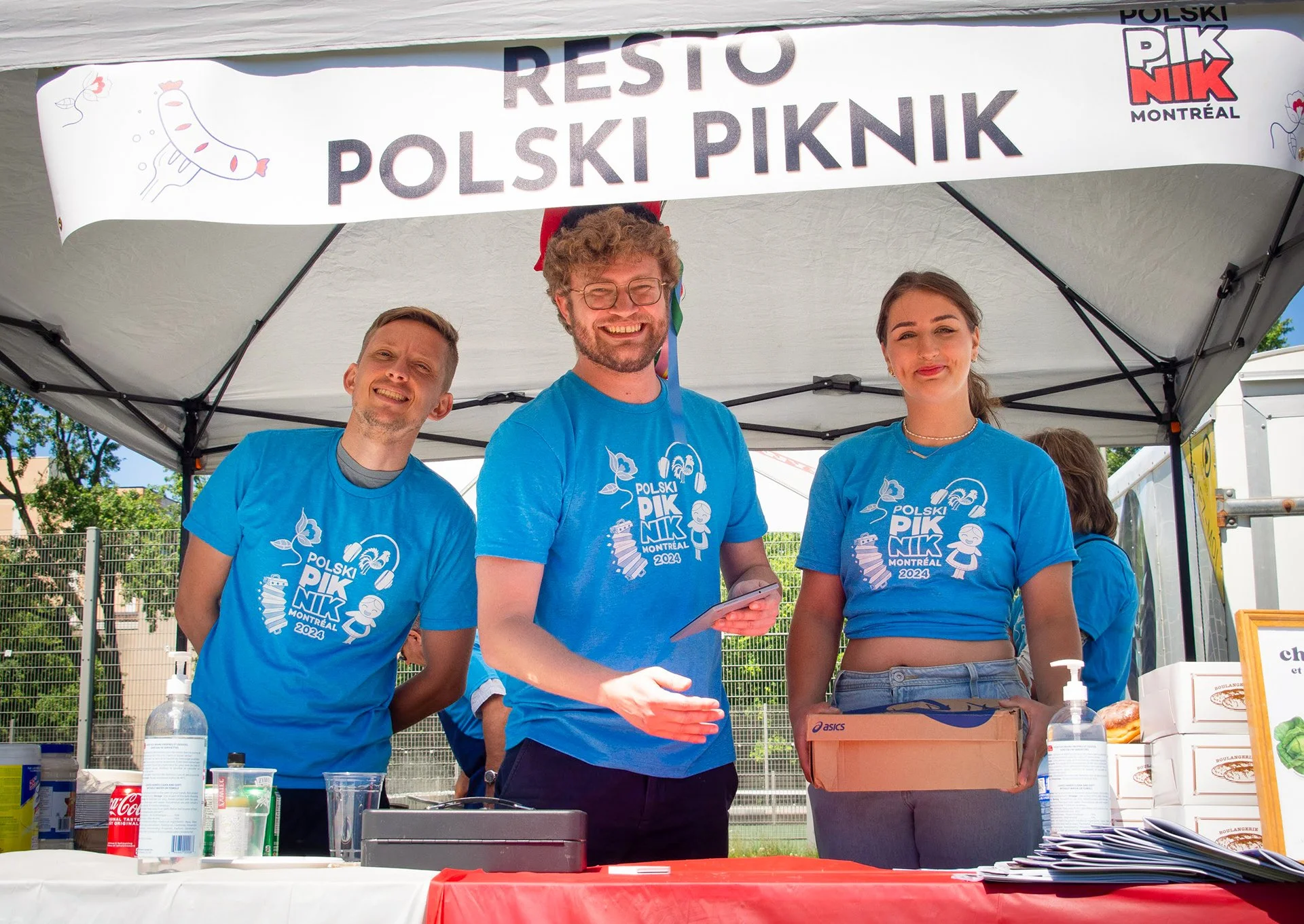 Four people standing behind a table at an outdoor booth, wearing blue t-shirts with a logo that reads 'Polski Piknik Montreal 2024.' One man in the center is smiling and holding a phone, and a woman to his right is holding a box. There are supplies on the table, and a banner above them reads 'Rest Polski Piknik' with a sausage illustration and event logos.