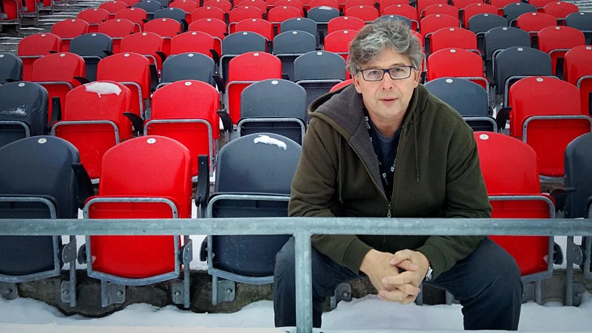 A man with glasses, gray hair, and a green jacket sitting and crouching in front of empty red and gray stadium seats with snow on the ground.