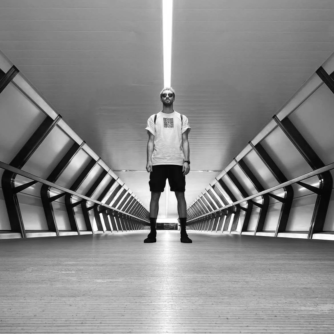 A man standing in the middle of an empty, modern, underground tunnel or corridor with metallic railings on both sides, wearing sunglasses, a T-shirt, shorts, and sneakers, in black and white.