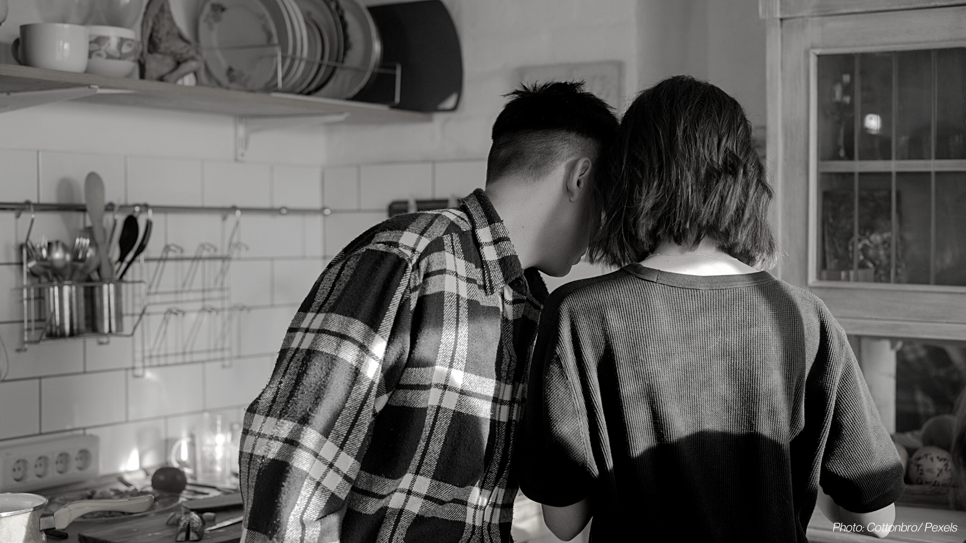 A black and white photo, taken from behind, of a couple cooking together in a kitchen