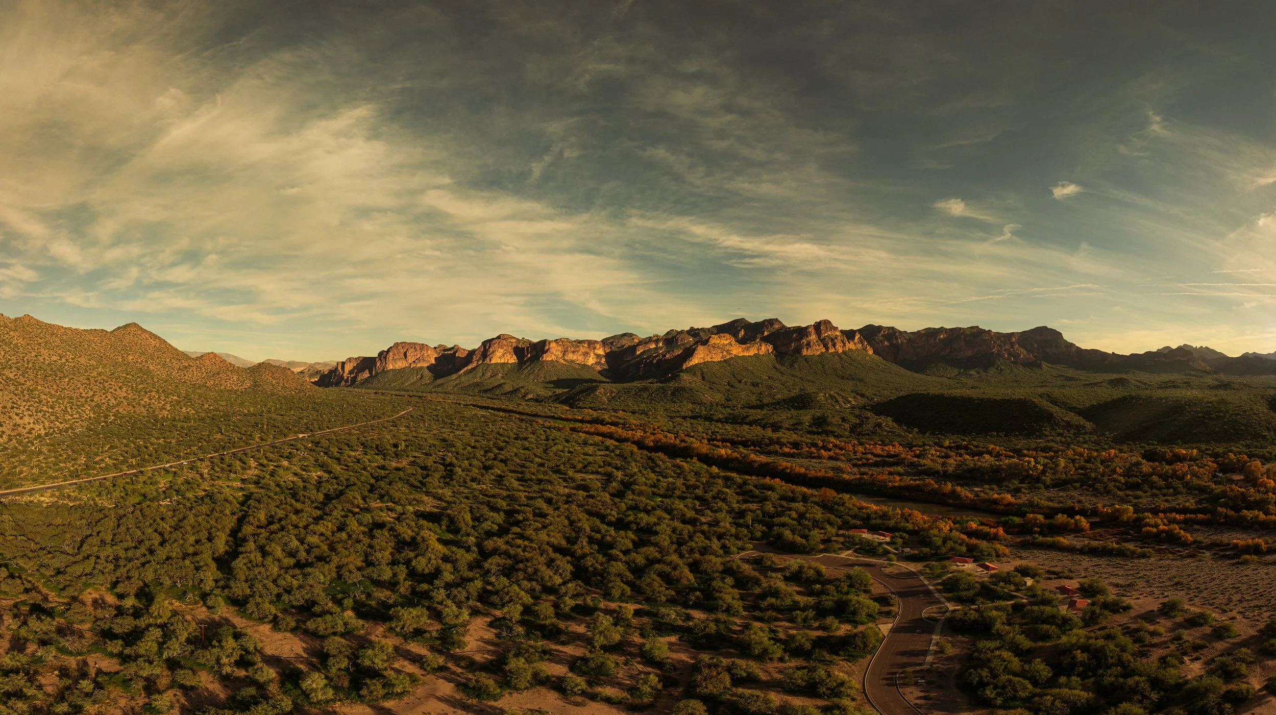 Sunset over a mountainous desert landscape with green shrubs and trees, and a winding road.