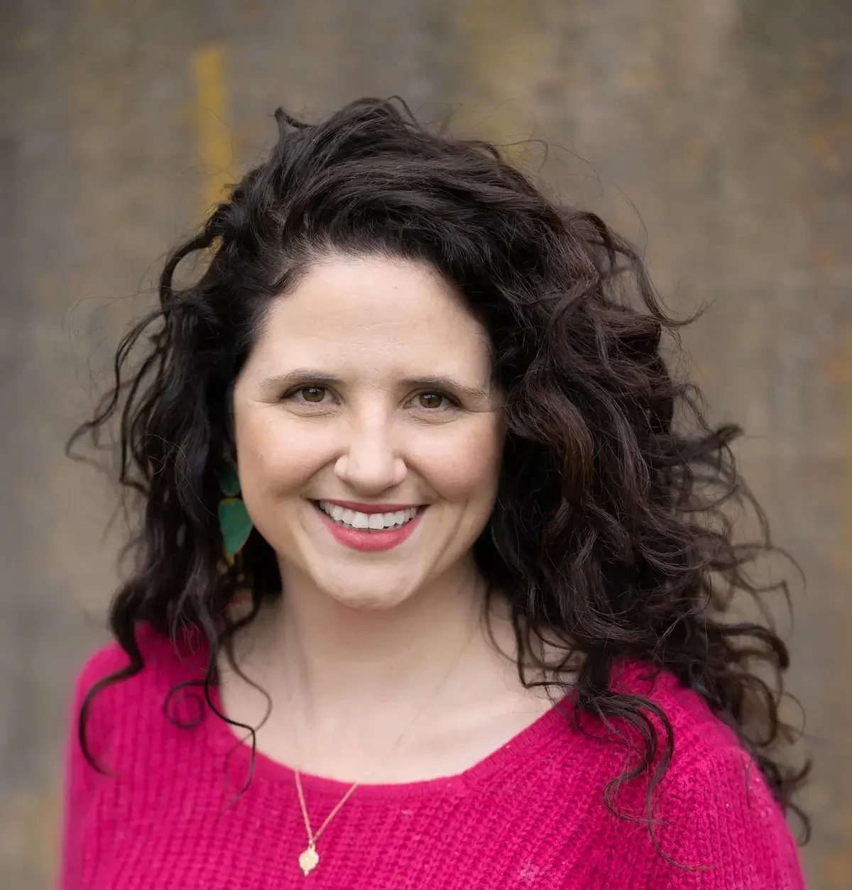 A woman with curly dark hair smiling, wearing a pink sweater, earrings, and a necklace, standing outdoors with a blurred background.