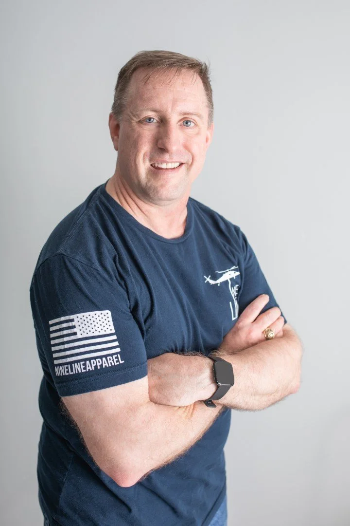 A smiling man with short light brown hair wearing a navy blue t-shirt with a helicopter graphic and the word "THE" on it, standing with his arms crossed, against a plain white background.