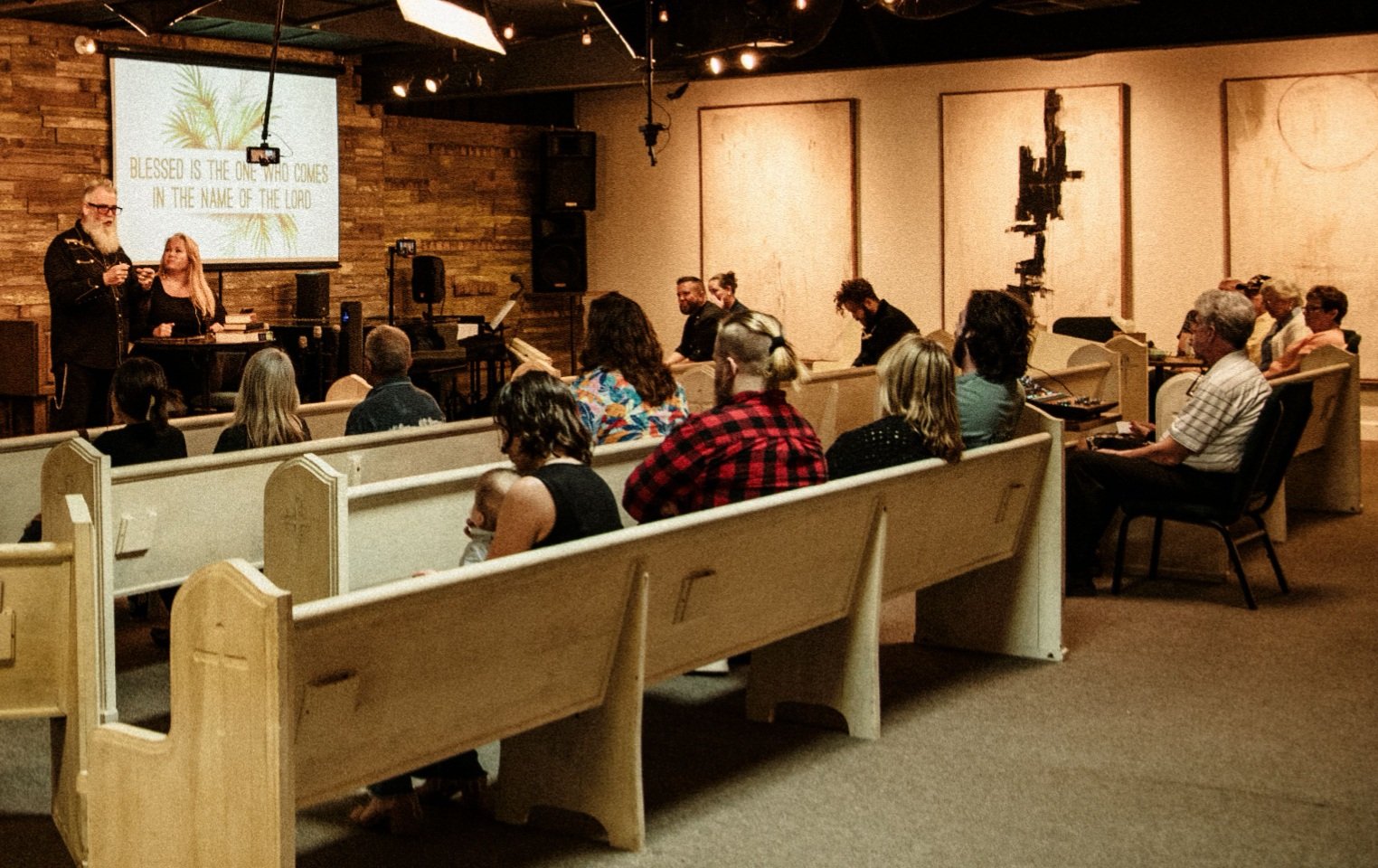 A church service with congregation members seated in pews, focusing on a speaker and a screen displaying the message 'Blessed is the one who comes in the name of the Lord.' The room has a warm, rustic interior with wooden panels and contemporary artw