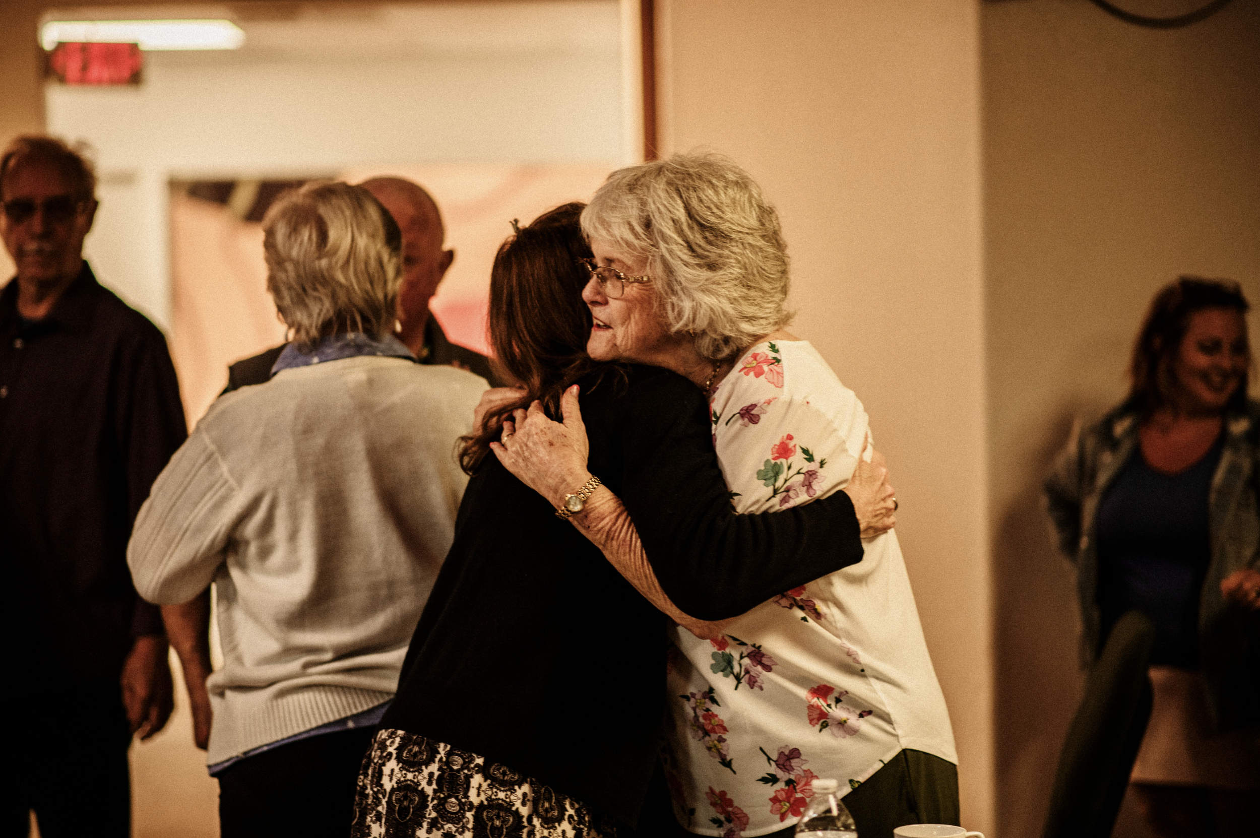 A group of people in church community, with focus on two women hugging, one wearing a floral blouse and glasses. Others in the background are conversing and smiling.