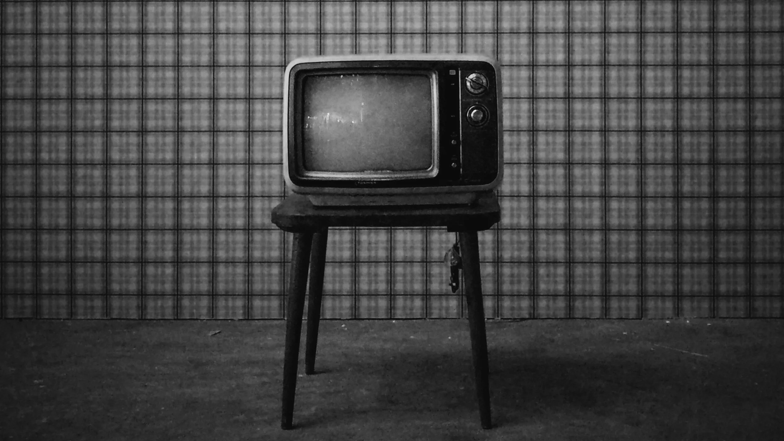 Black and white photograph of an old television set on a stand in front of a tiled wall, with a dark floor