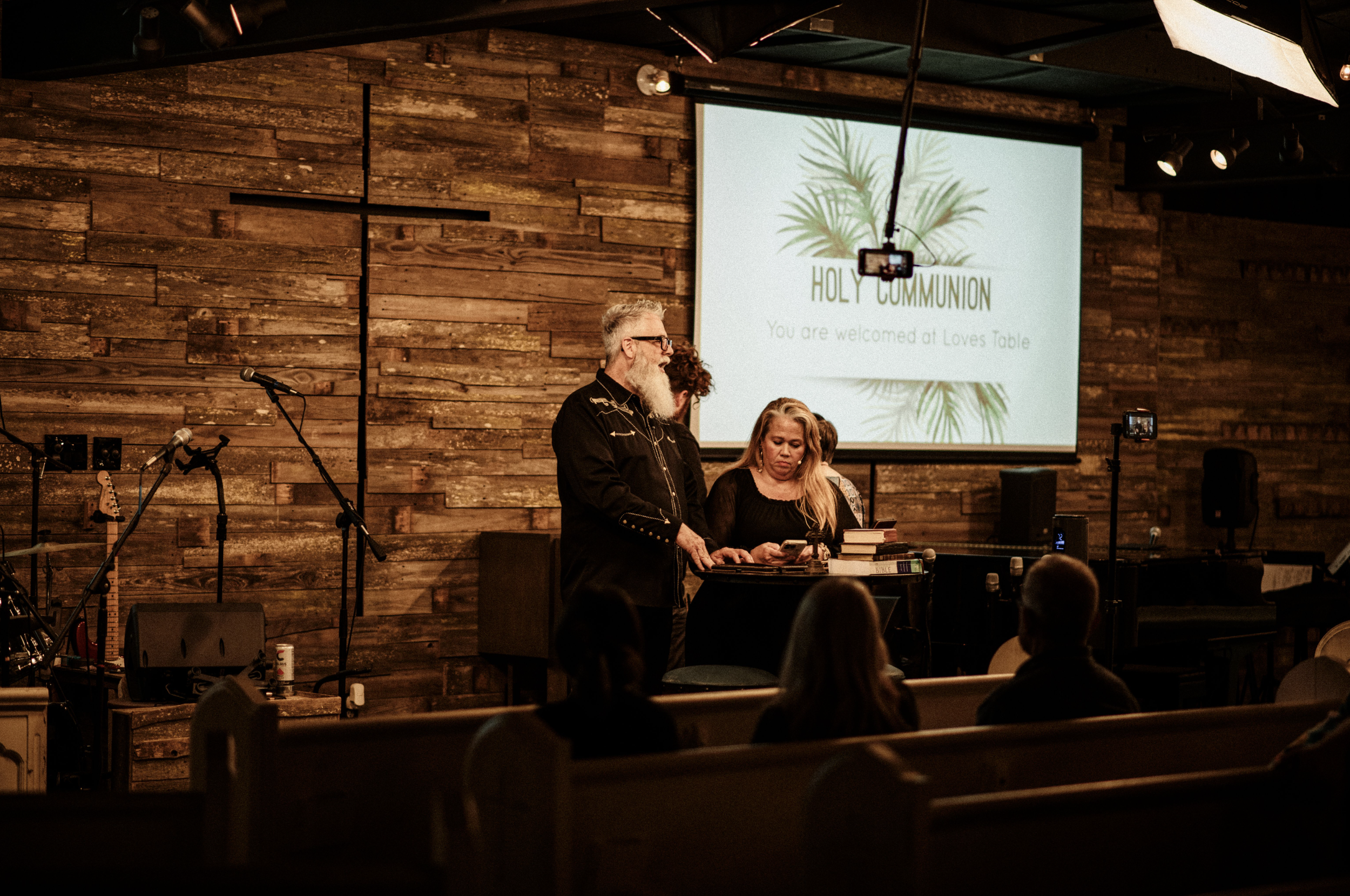 A church interior in Ocala Florida with a wooden cross, microphone stands, a guitar, and man and woman pastor near a pulpit. A screen displays "Holy Communion You are welcomed at Loves Table."