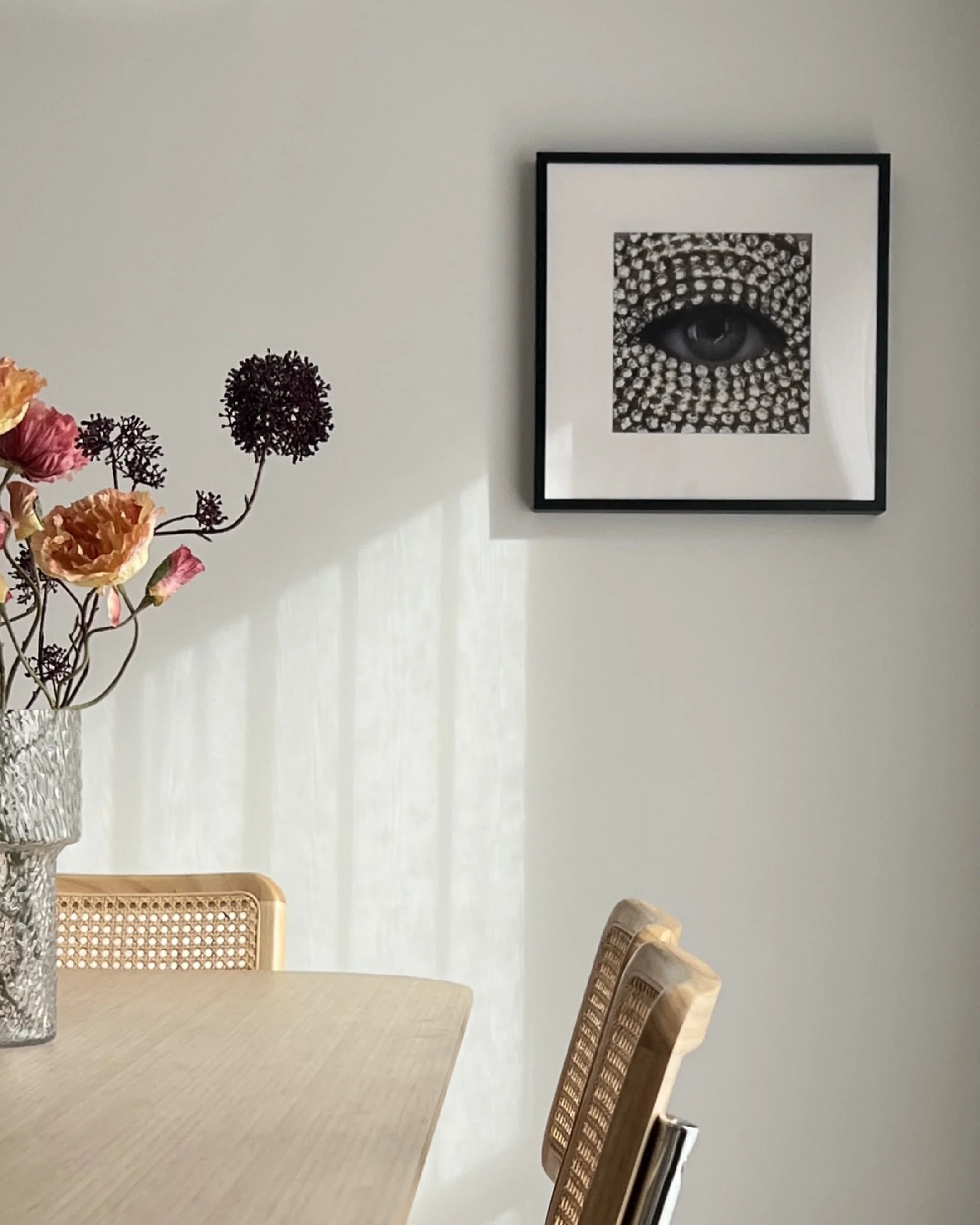 A dining room with a table, two woven chairs, a floral vase with pink and orange flowers, a piece of wall art featuring an eye surrounded by small diamanté stones, and a white wall.