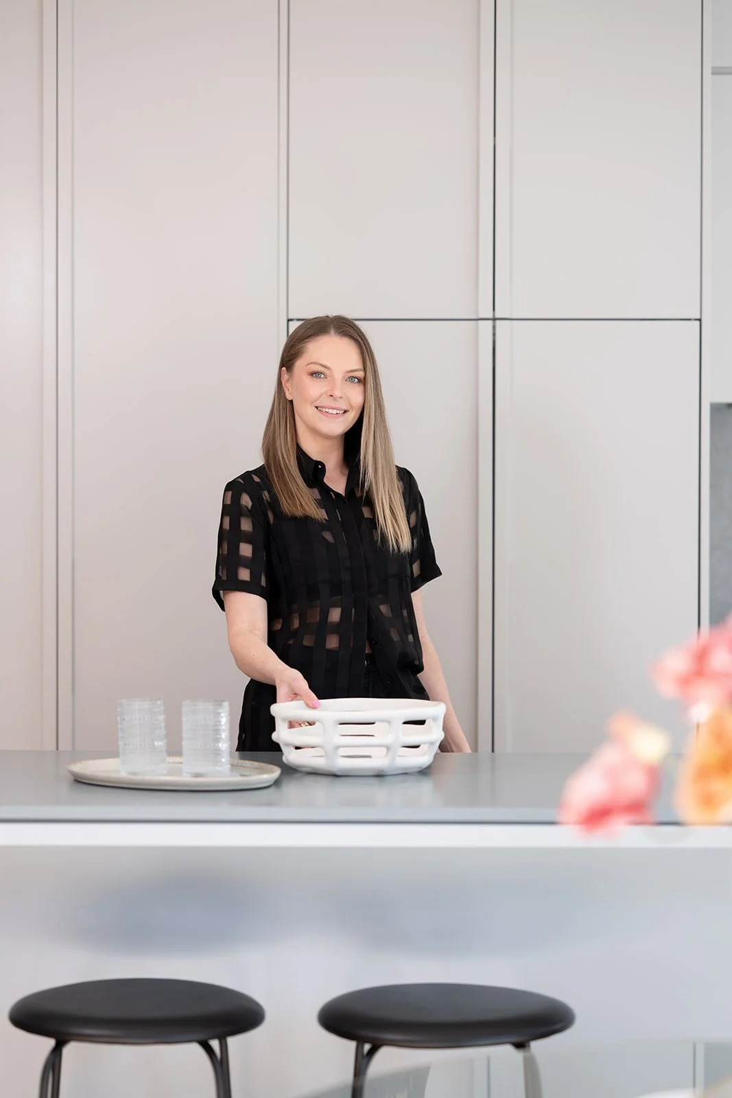 A smiling woman with straight, shoulder-length hair holding a white basket in a modern kitchen with white cabinets and a gray countertop.