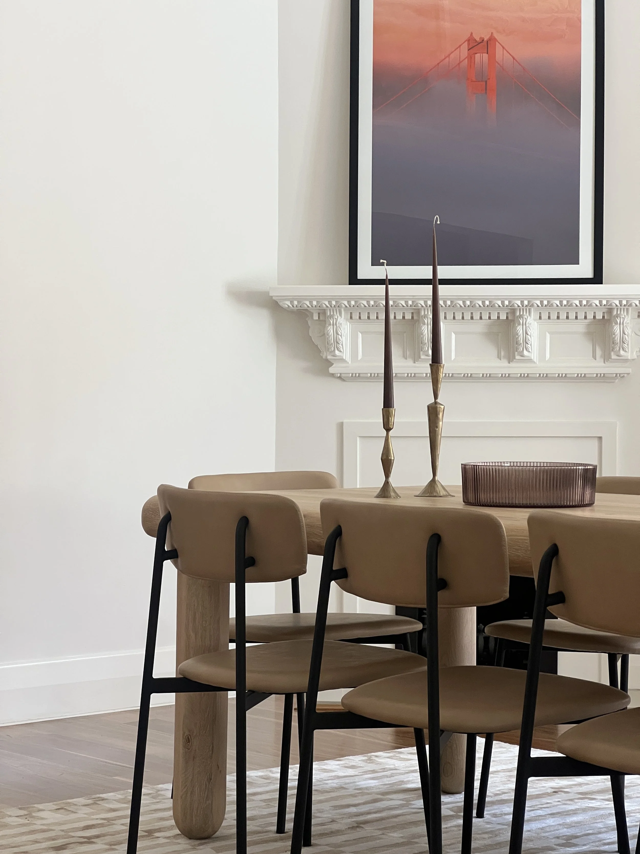 Interior of a dining room with a wooden table, six tan chairs with black legs, and decorative candles on the table. A framed picture of the Golden Gate Bridge at sunset is hung above a white fireplace mantel.