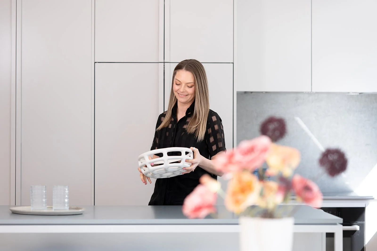 A woman with shoulder-length blonde hair smiling while holding a white basket in a modern kitchen with white cabinetry. There is a vase of pink flowers on the kitchen island and a plate with glasses.