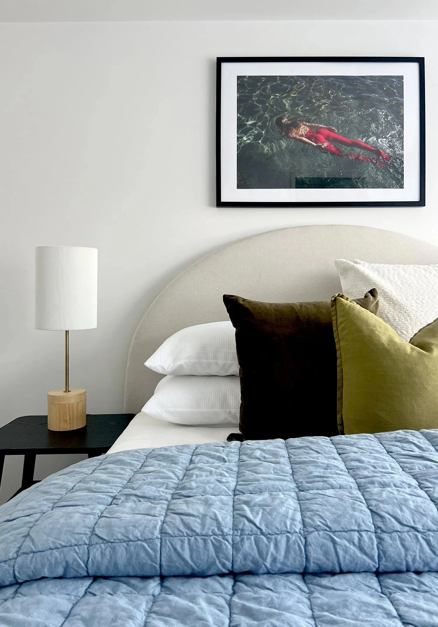 A neatly made bed with a light grey headboard, a white pillow, and several decorative pillows in black, green, and white. A black nightstand with a lamp featuring a white shade and wooden base is beside the bed. Above the bed, there is a framed photograph of a person swimming in water, wearing red pants, and the wall is plain white.