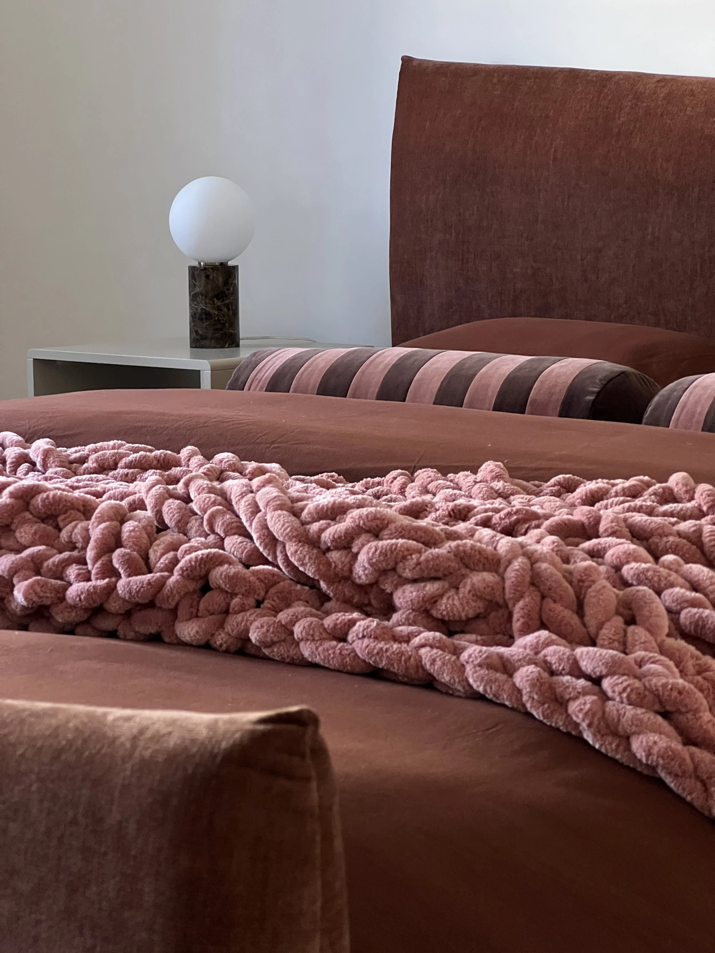 Close-up view of a pink plush blanket on a bed with pink and brown pillows, a pink headboard, and a white nightstand with a modern lamp in the background.