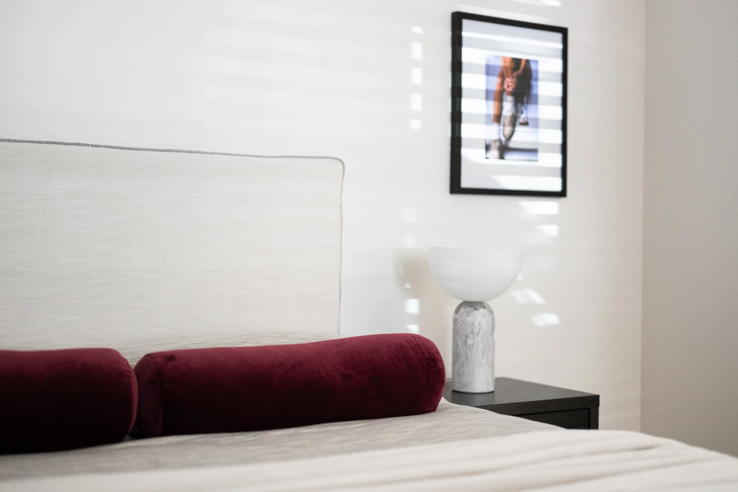 A contemporary bedroom with a light gray upholstered bed, a red velvet bolster cushion, a black timber side table with a modern marble a lamp, and a black-framed artwork on the wall featuring horizontal light stripes.