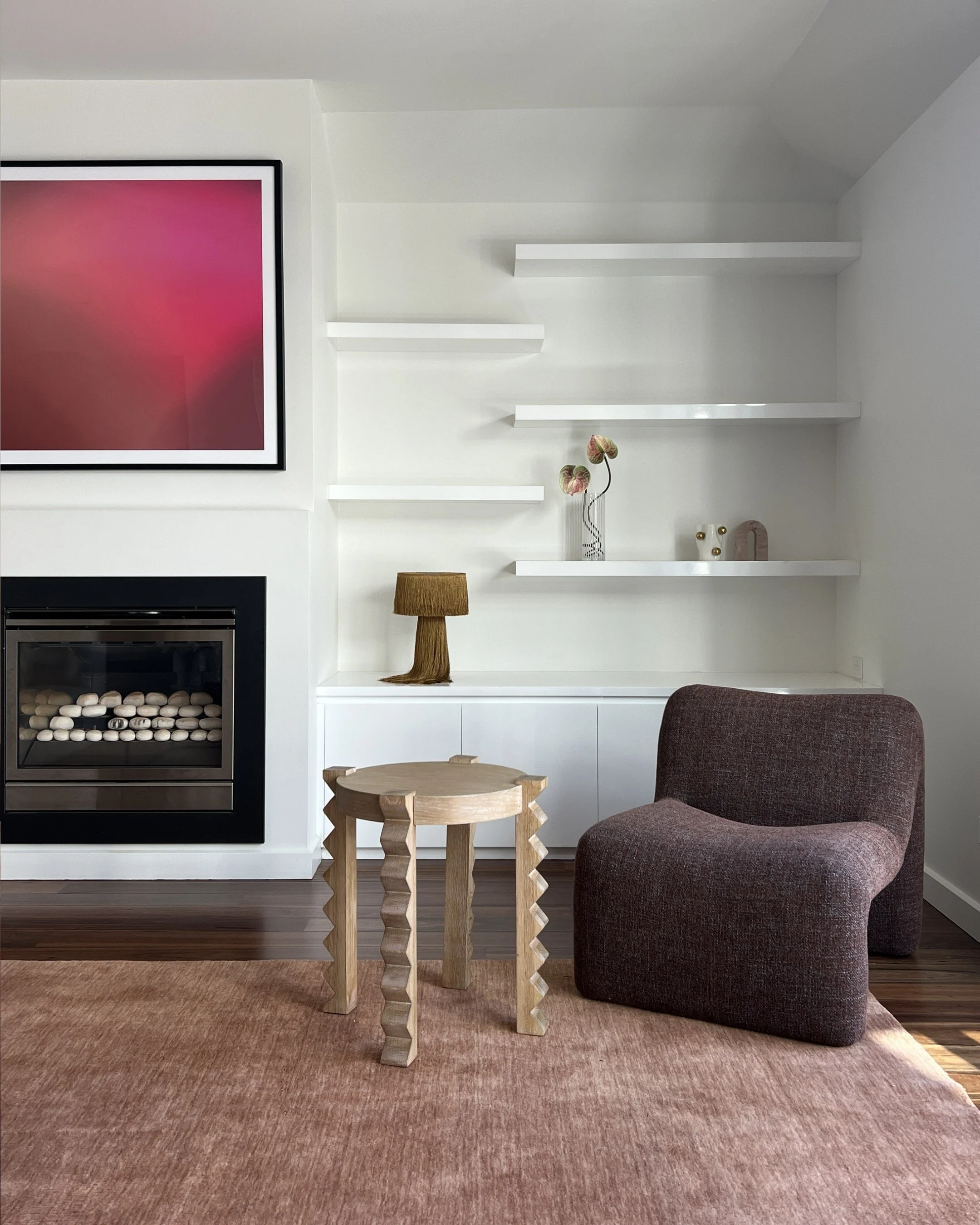 Modern living room with a burgundy chair, small wooden table, fireplace, and white wall with built-in shelves and decorative items.