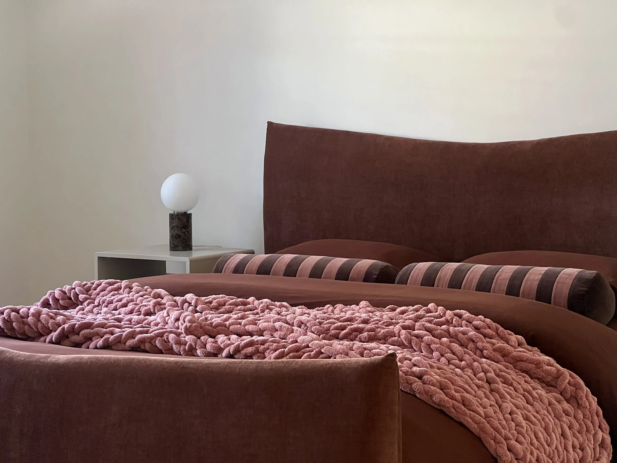 A bedroom with a maroon upholstered headboard, a bedside table with a lamp, and a textured pink blanket on the bed.