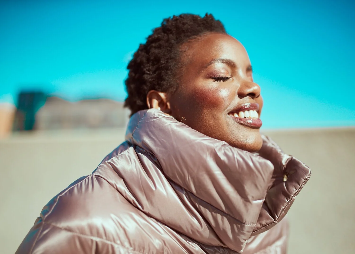A woman with short curly hair smiling with her eyes closed, wearing a shiny beige puffer jacket, outdoors under a clear blue sky.