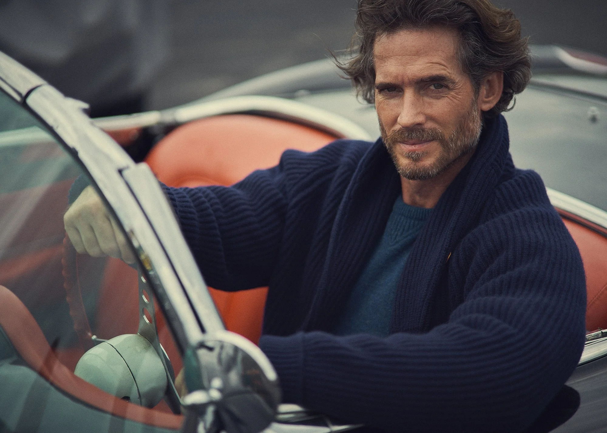 A man with brown hair and a beard sitting in a vintage car with orange leather seats, wearing a navy blue sweater, looking at the camera.