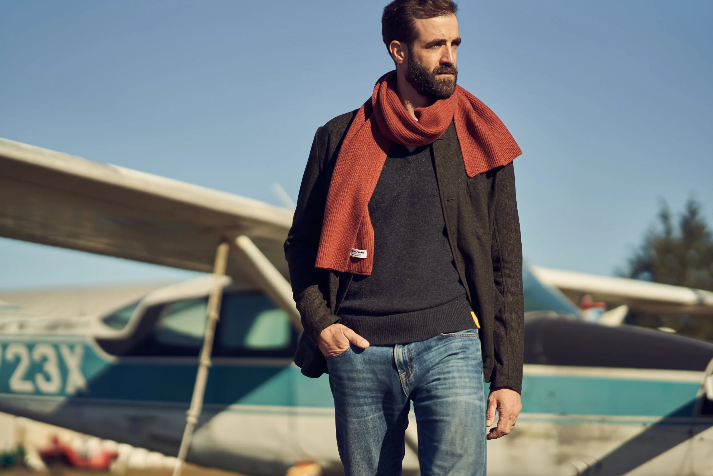 A man with a beard and brown hair dressed in a black jacket, black sweater, blue jeans, and a red scarf standing near small airplanes outdoors on a clear day.