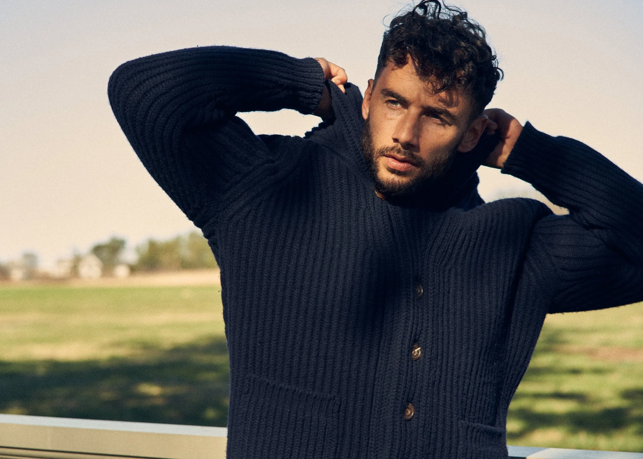 A man with curly hair and a beard wearing a dark knit sweater outdoors, adjusting his hoodie with both hands, under a cloudy sky.