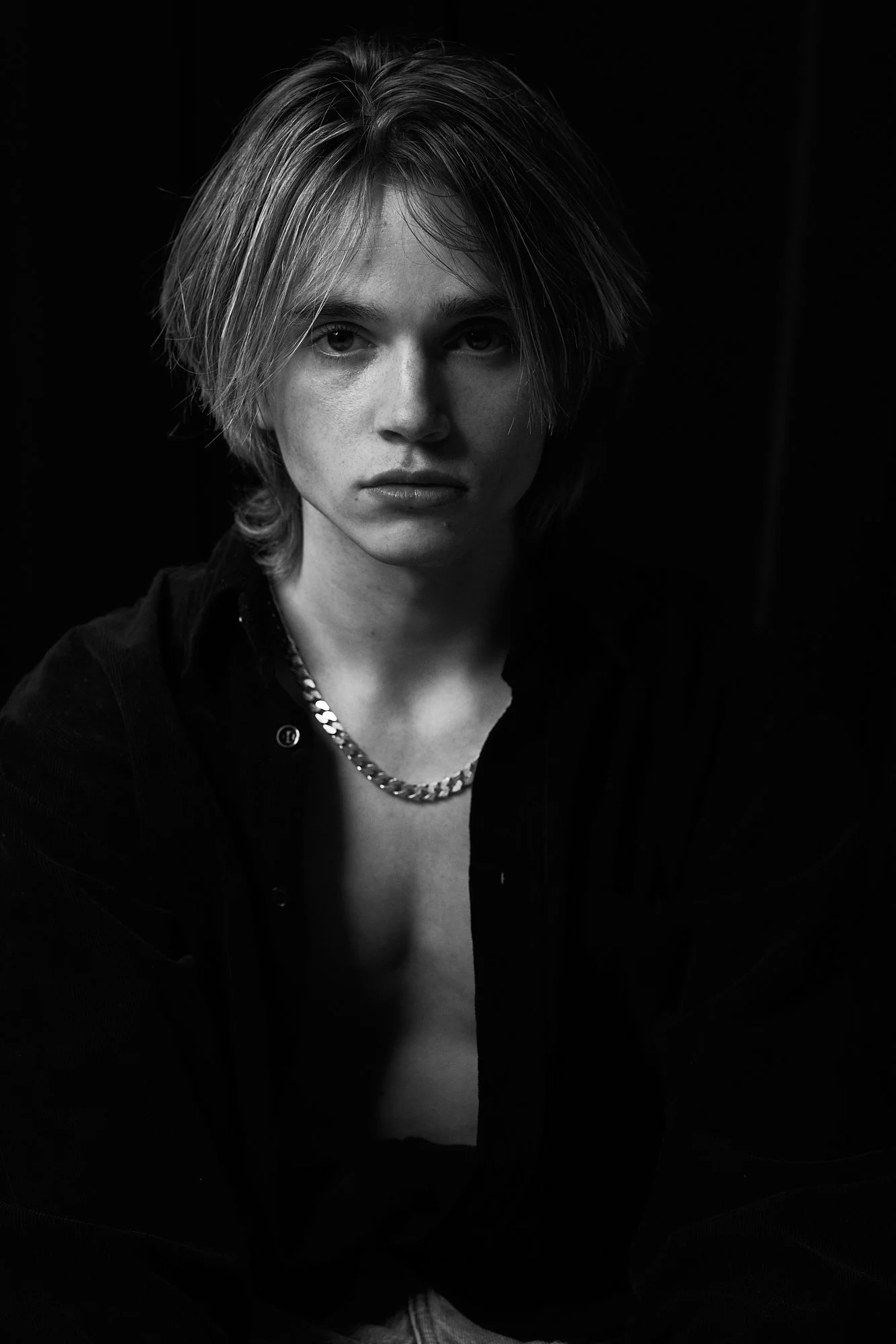 Black and white photo of a young woman with short, layered hair wearing a dark shirt and a chain necklace, looking directly at the camera.