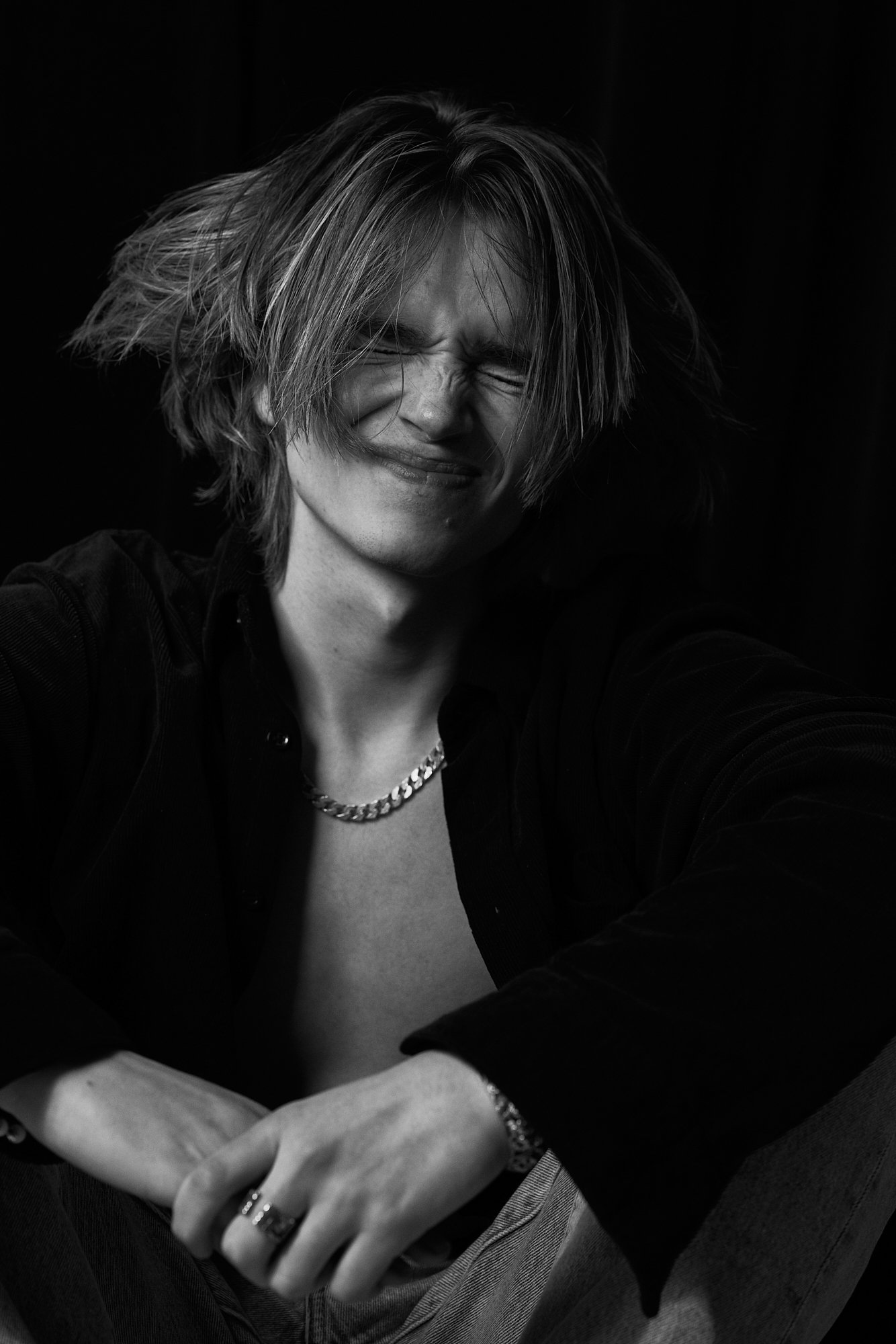 Black and white photo of a person with shoulder-length hair, eyes closed, smiling, wearing a chain necklace, rings, a bracelet, and a black shirt