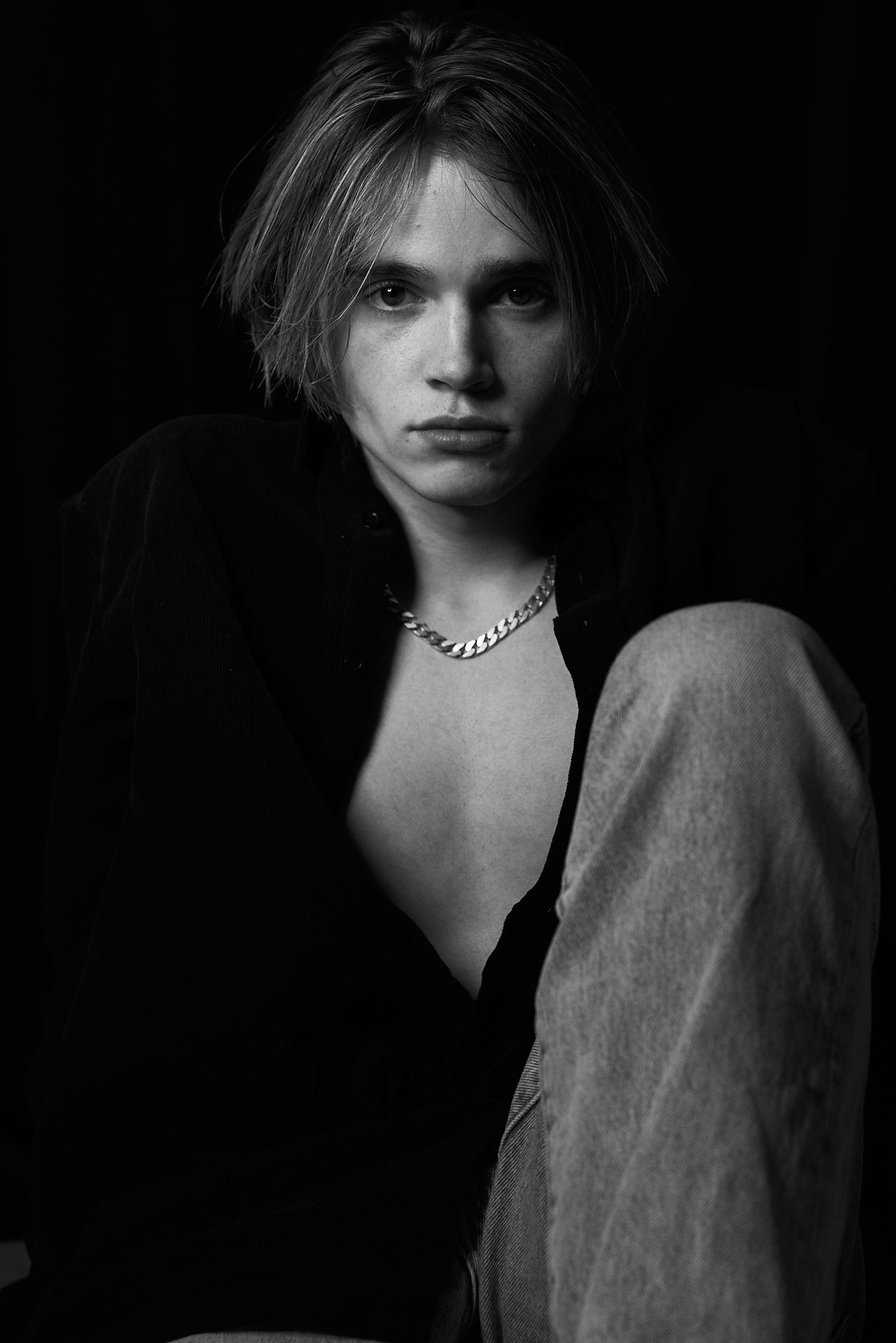 Black and white portrait of a young woman with short, tousled hair, wearing a chain necklace, a dark open shirt, and light-colored pants, sitting against a dark background.