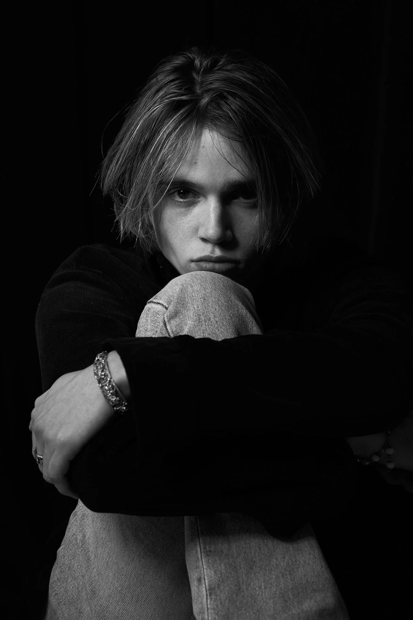 Black and white portrait of a young person with short, disheveled hair, sitting with one knee up and arms wrapped around it, looking directly at the camera with an intense expression.