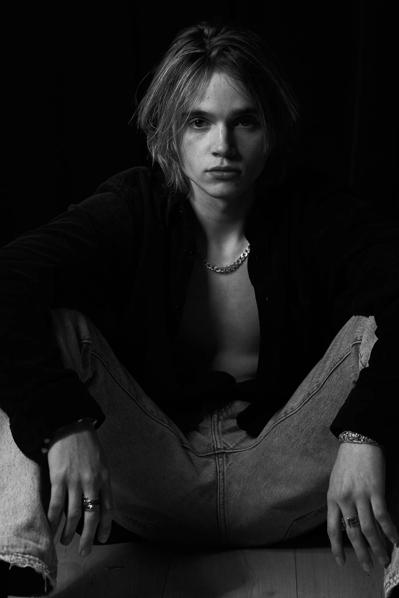 A black and white portrait of a person with shoulder-length hair sitting with legs apart, wearing a dark shirt, light jeans, and jewelry, looking directly at the camera.