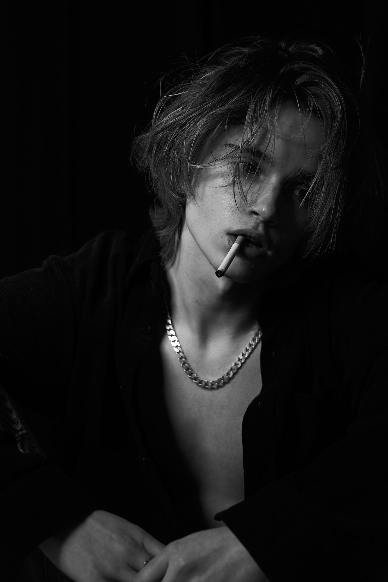 A young person with messy hair, wearing a chain necklace and dark clothing, holds a cigarette in their mouth, in a black and white portrait.