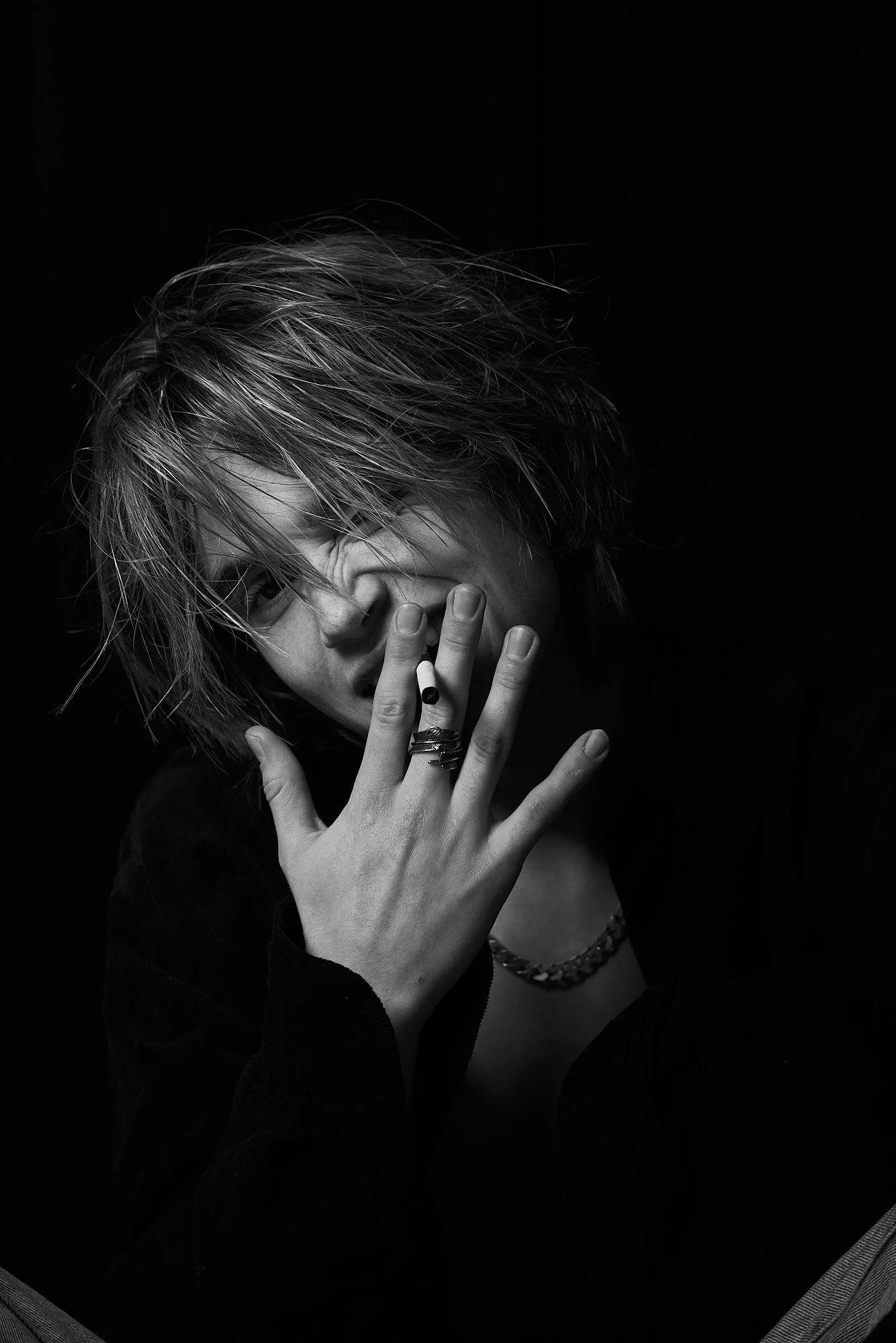 Black and white photo of a person with messy hair, partially covering one eye, holding a cigarette near their mouth, wearing rings, a necklace, and a dark shirt, with a dark background.