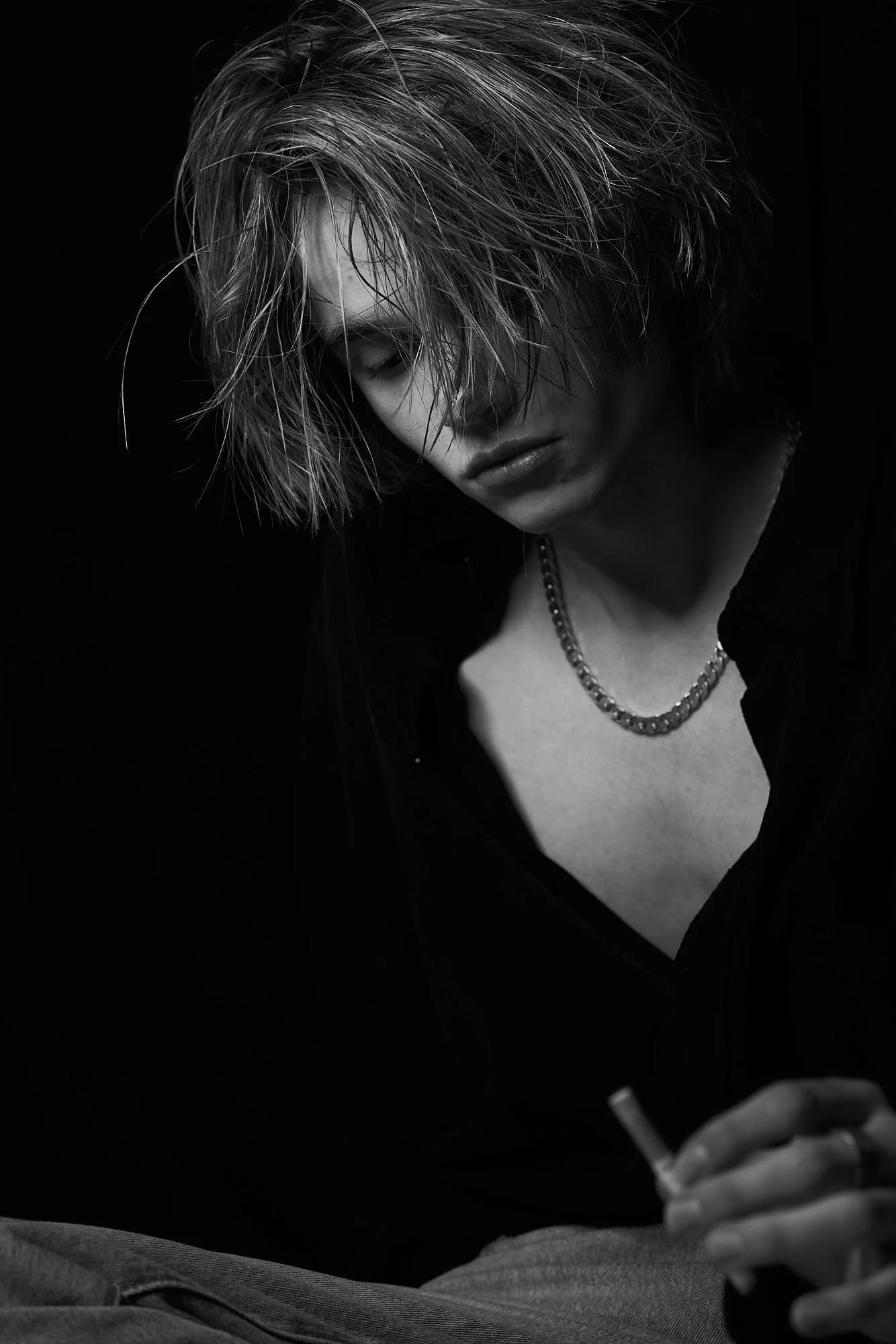 Black and white photo of a woman with messy, short hair, wearing a necklace, and a dark shirt, looking down and holding a lit cigarette.