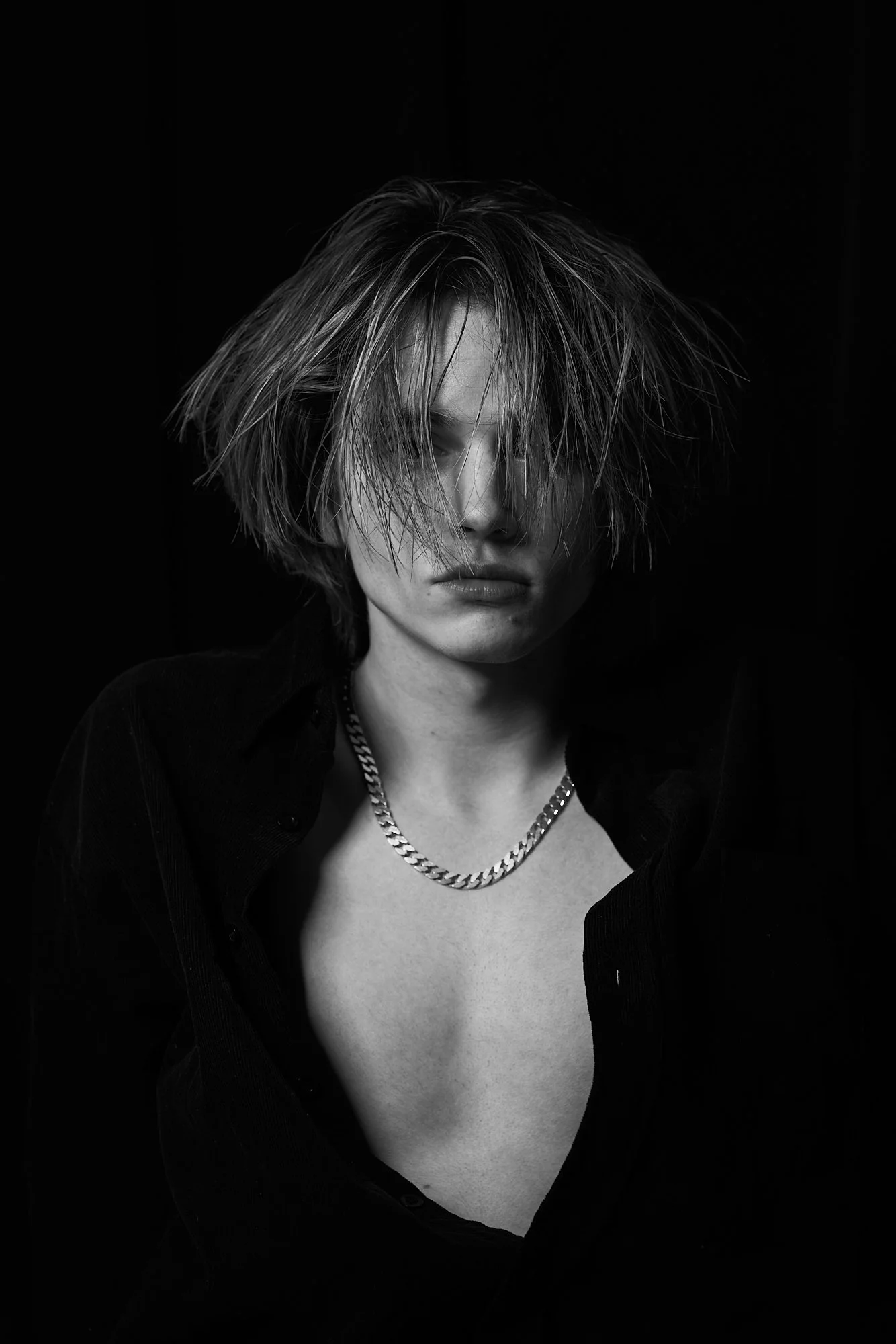 Black and white portrait of a woman with messy short hair, wearing an open dark shirt and a chain necklace, looking directly at the camera with a serious expression.