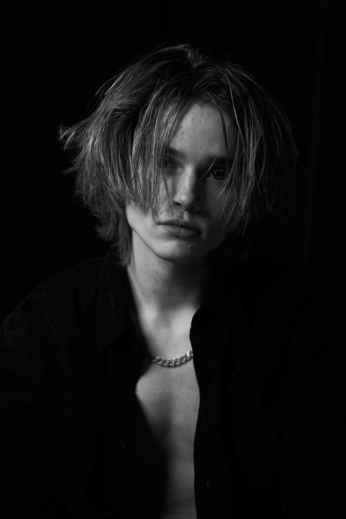 Black and white portrait of a young person with tousled hair, wearing a dark shirt and chain necklace, looking at the camera with an intense expression.