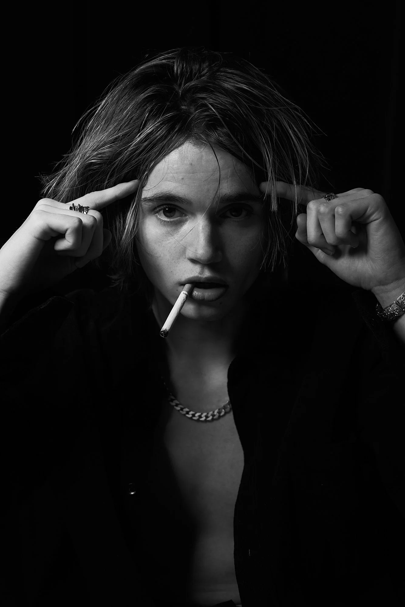 Black and white portrait of a young woman with short hair, finger pointing to her temple, with a cigarette in her mouth, wearing a chain necklace and rings.